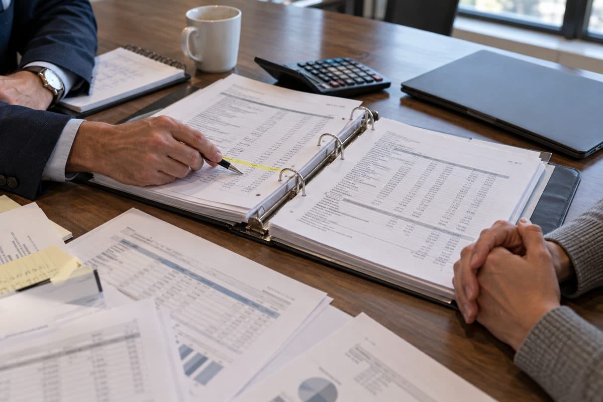Two people review a binder of vendor invoices with one line highlighted